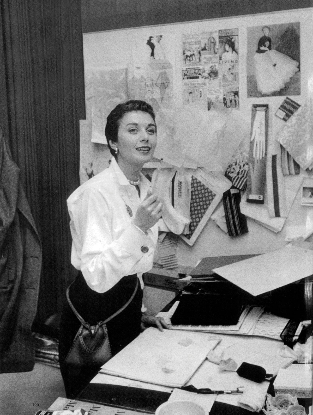 Anne Klein in white blazer seated at desk with fashion design sketches and fabric swatches pinned to wall behind her.