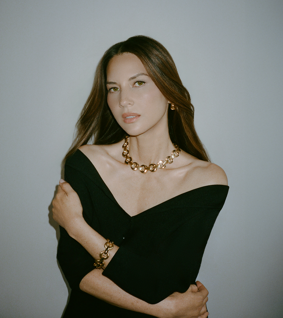 Woman in black off-shoulder dress with gold chain necklace and matching bracelet, against neutral background.