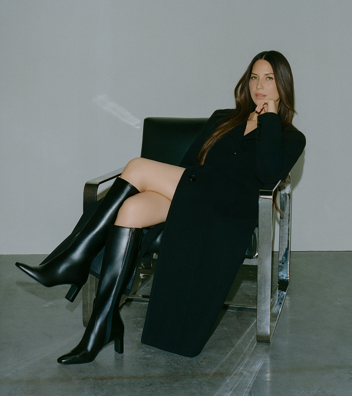 Woman in black blazer and black boots seated in modern chair against gray background