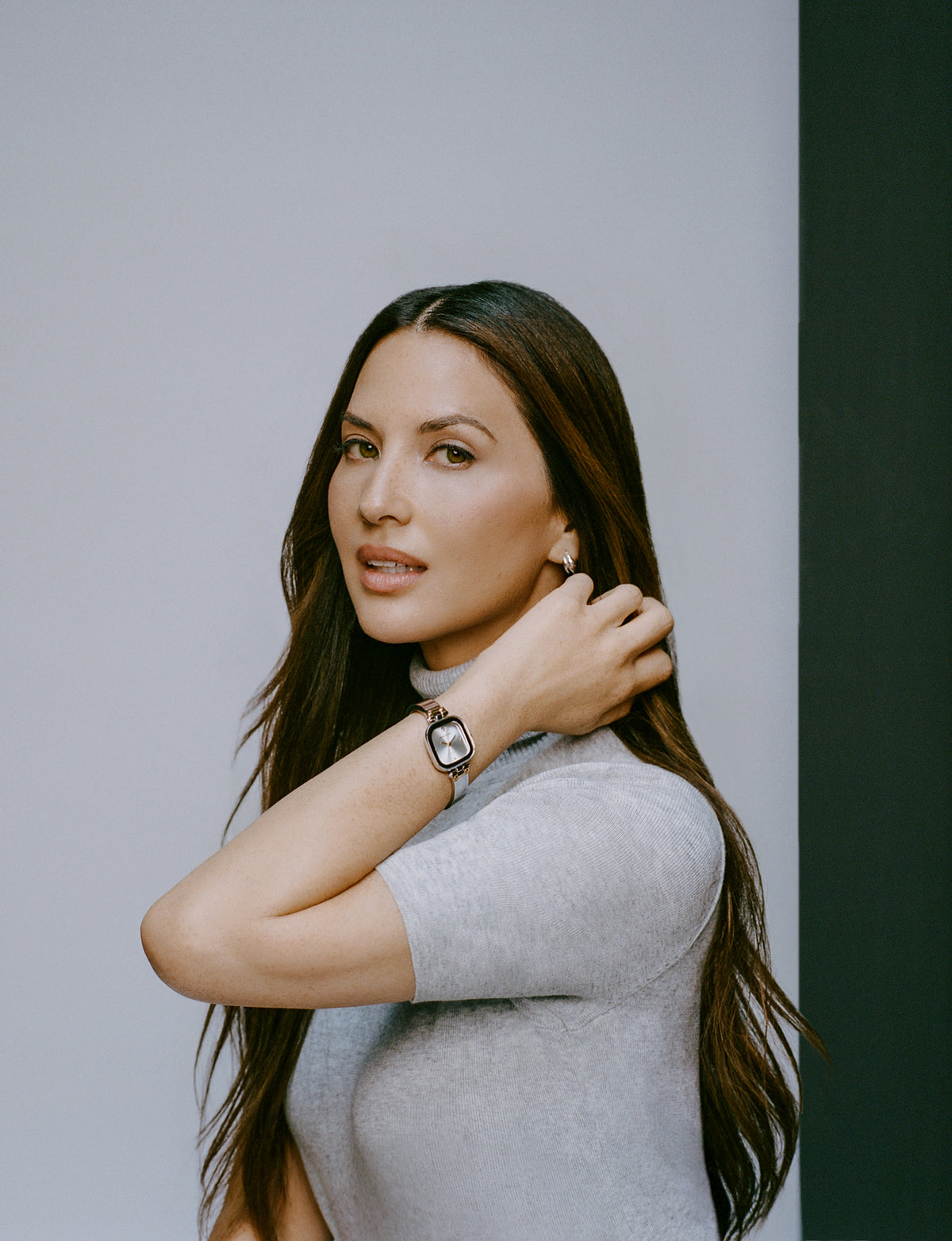 Woman in light gray turtleneck wearing rectangular watch, looking over shoulder against two-tone background.