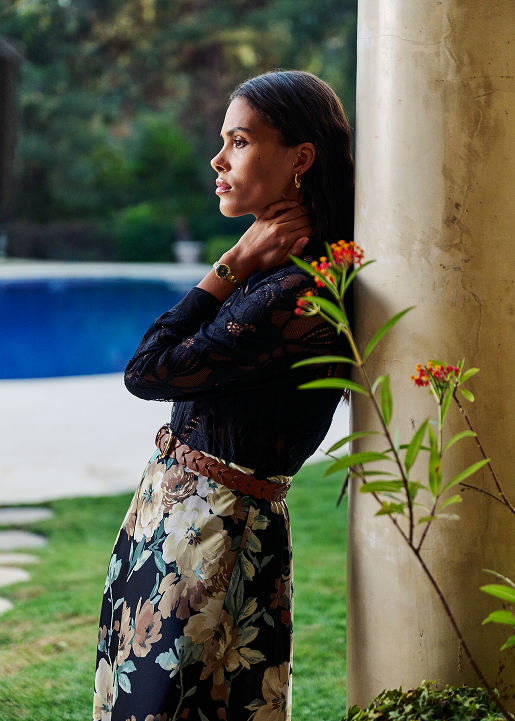 Woman in black jacket and floral skirt standing against column by pool, small red flowers visible nearby