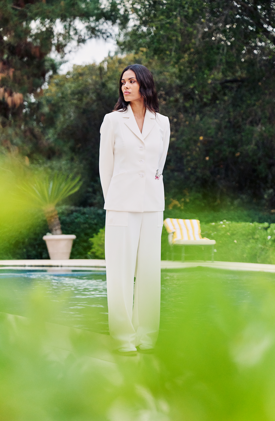 Woman seated on ornate chair in tailored cream blazer and trousers, wearing jewelry, on manicured lawn with formal balustrade and trees in background