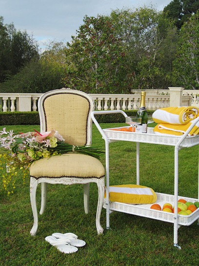 Woman seated in cream upholstered chair on lawn, white side table with yellow accents and fresh citrus fruits nearby, manicured garden and balustrade in background.