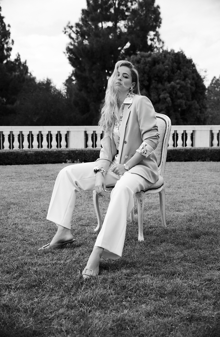 Woman seated on ornate chair in tailored cream blazer and wide-leg trousers, formal garden setting with manicured hedge and balustrade behind