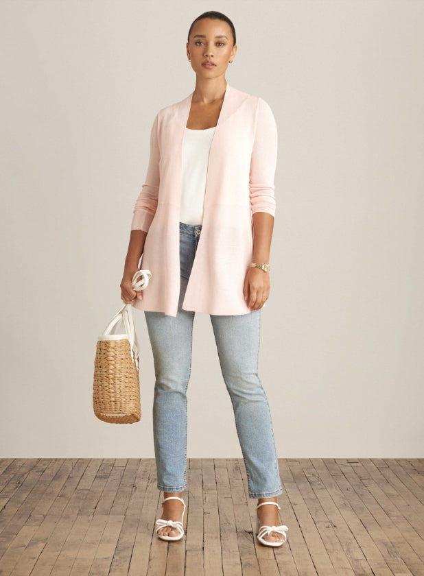 Model in blush pink cardigan, light wash jeans and white sandals holding a straw tote bag