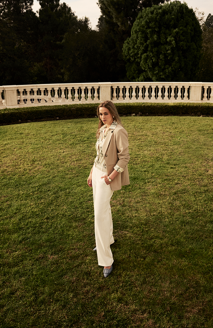 Woman in tan blazer and cream pants standing on grass with formal hedge and topiary in background