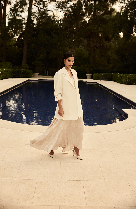Woman in cream blazer and beige pleated skirt standing on pool deck at dusk