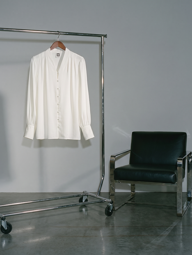 Cream button-front blouse on hanger with puffed sleeves, displayed on chrome clothing rack next to dark green chair.